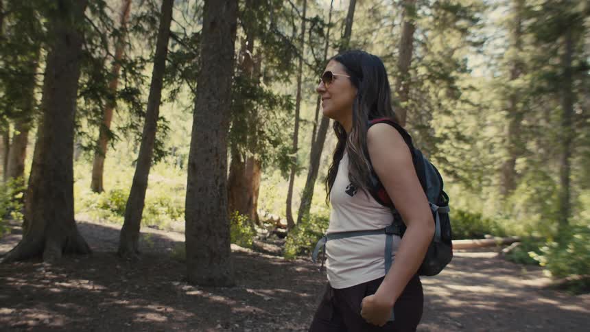 A woman walks through a sunlit forest wearing a backpack and sunglasses