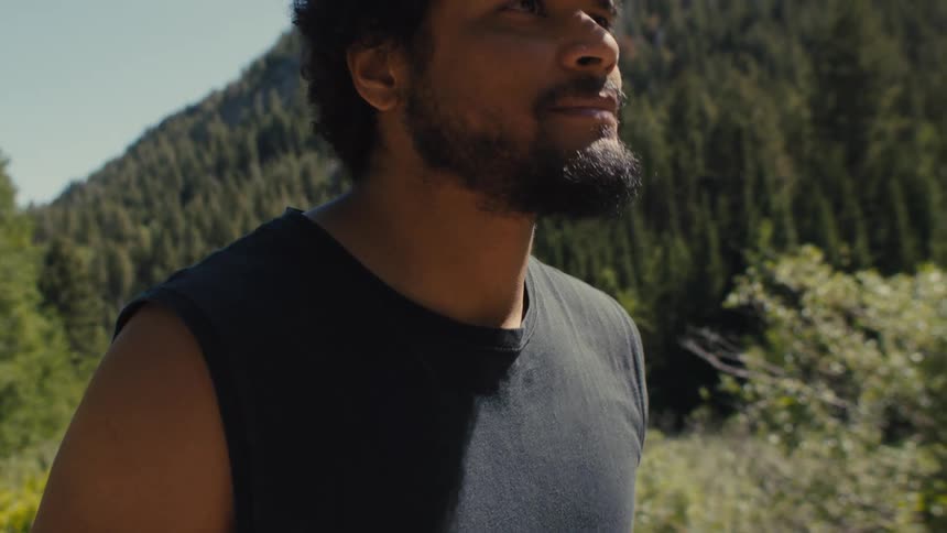 A man walks outdoors in a forested mountain area under bright daylight