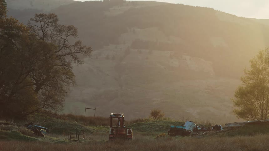 An excavator sits in a grassy rural field surrounded by scattered debris at sunset