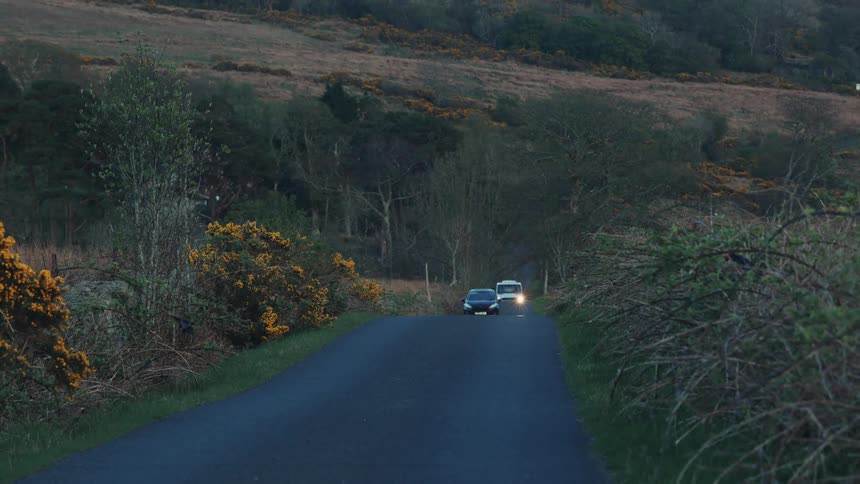 Two vehicles travel a quiet rural road through hilly bush lined countryside
