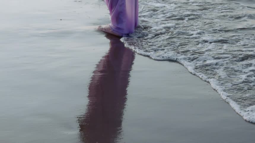 Person wearing filmy pants stands on the reflective sand as the surf flows onto the beach