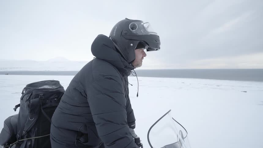 A man puts on a helmet and climbs onto a snowmobile in the snowy wilderness