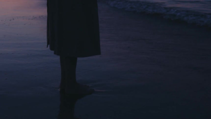 Person in a long garment stands barefoot at the edge of a twilight shore with soft waves lapping against wet sand under a fading sky