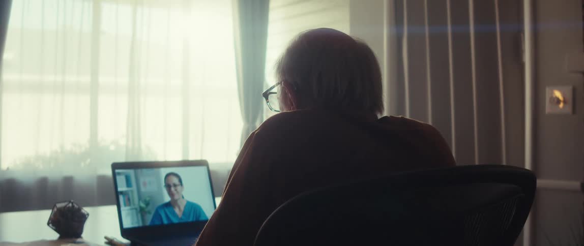 Elderly man sitting at a table facing a laptop showing a female nurse