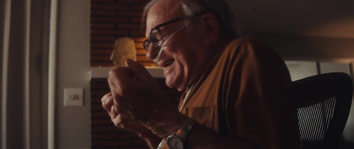 An elderly man with a nasal cannula on his nose sits behind a desk in his living room at midday looking and acting angry while speaking and gesturing with his hands