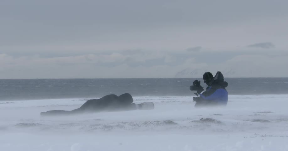 Two people with camera equipment sit near the snowy shore