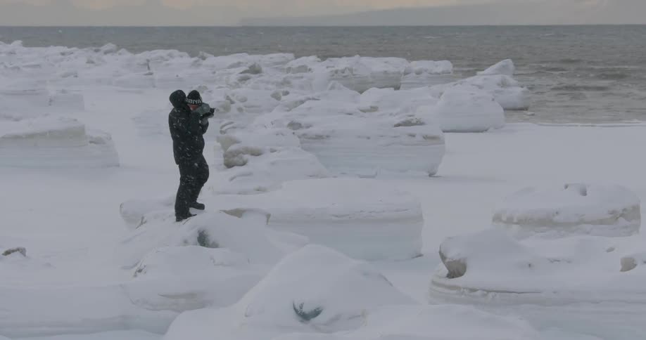 A man stands on rocks and ice taking photographs in the snow