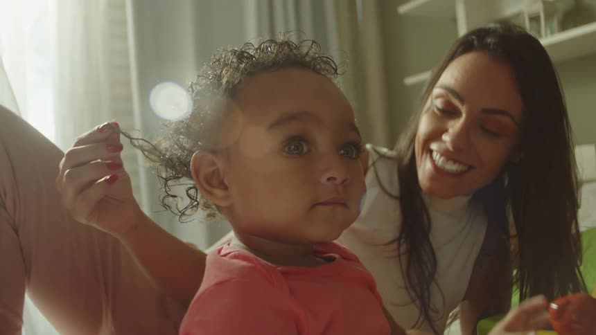 Child in a pink shirt plays with a yellow toy as a smiling woman gently caresses her hair in a sunlit room
