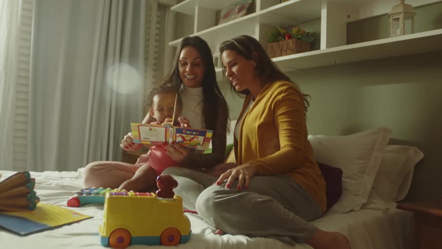 Mother and grandmother read a book to a toddler while all three sit on a bed