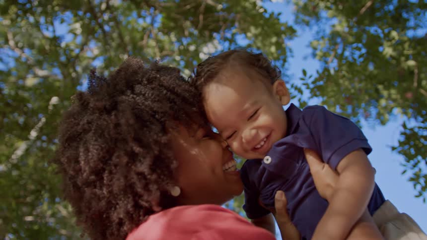 A woman lifts a laughing child into the air under a canopy of green trees
