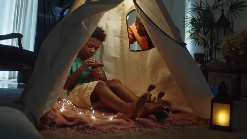 Boy playing inside a makeshift tent in the living room and his mother watching nearby