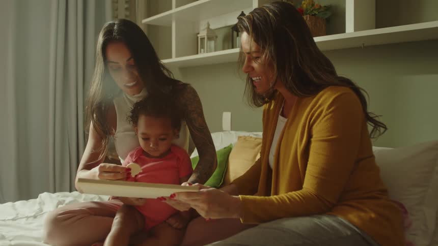 Two women and a baby sit on a bed looking at a book together