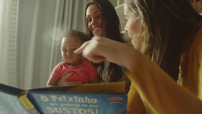 A mother holds her child while another woman reads from an open book to the child indoors