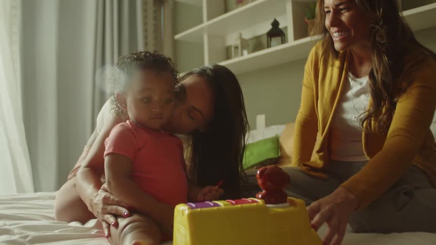 Two women joyfully engage with a toddler playing on a bed in a cozy room