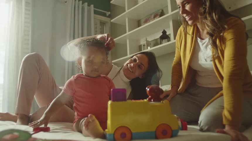 Toddler playing with a toy on the bed while two women interact joyfully nearby
