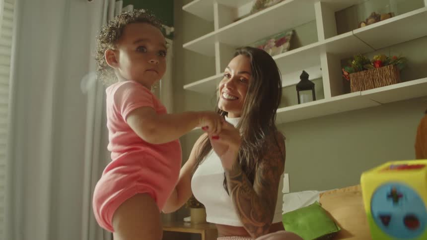 Two women smile while engaging with a baby wearing a pink bodysuit in a bright room with shelves in the background