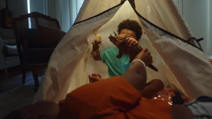Brothers make toy dinosaurs fight each other while one sits in a small tepee in the living room