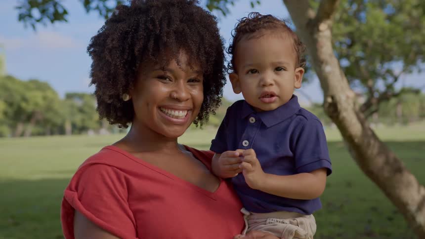 A smiling woman is holding a young child in a sunny park