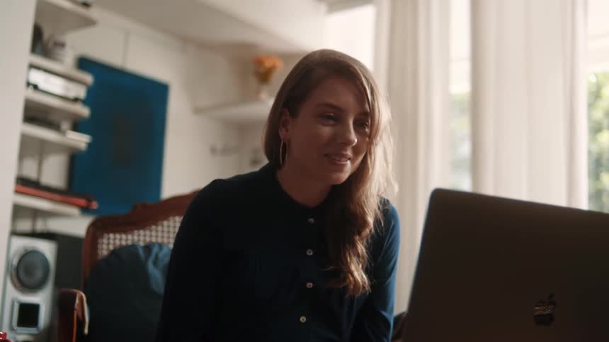 A woman is smiling while video chatting on a laptop in a cozy sunlit room