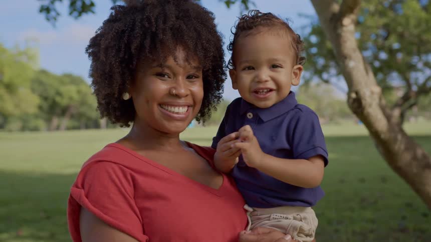 A black woman smiling while holding a small black child outdoors near a tree