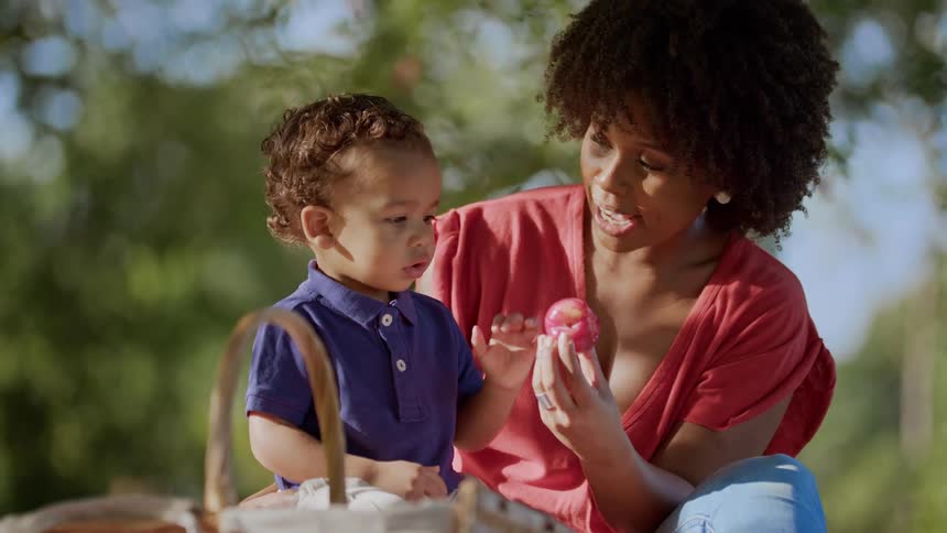 A woman in the park gives a fruit to her toddler while having a picnic around midday