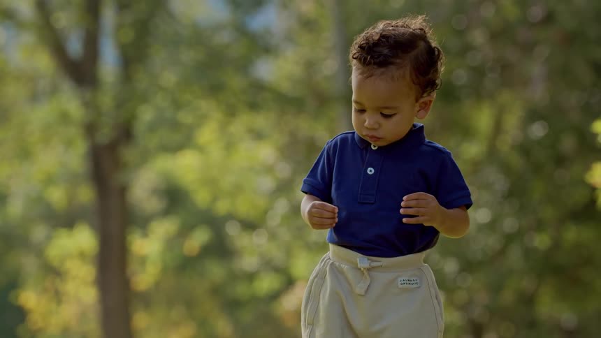 A toddler walks alone in a sunny park surrounded by trees