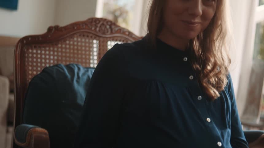 A pregnant woman seated on a chair indoor smiles at dawn