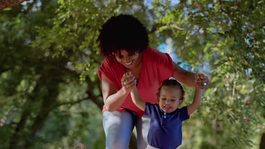 A smiling woman walks outdoors holding a happy toddlers hands under the trees