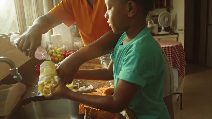 A woman and a child are washing dishes at a kitchen sink with sunlight streaming through a window