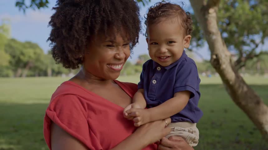 A smiling mother lovingly tickles her toddler son in a sunny park