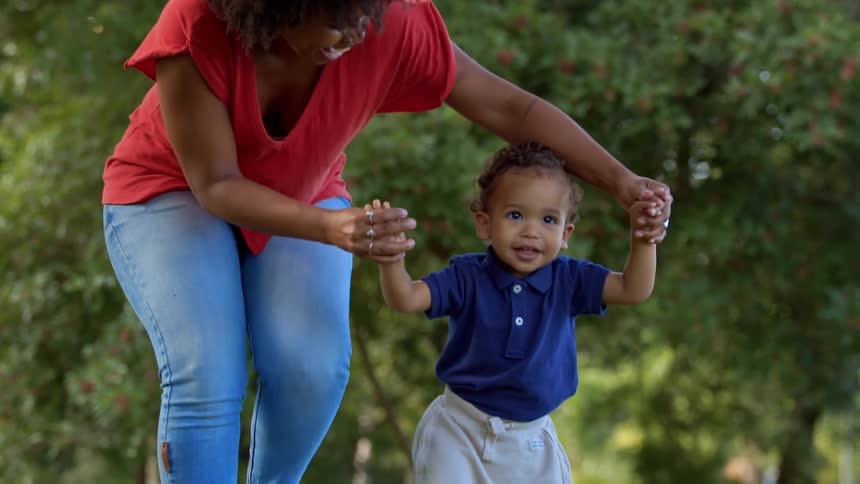 A joyful mother helps her toddler walk outdoors under leafy trees