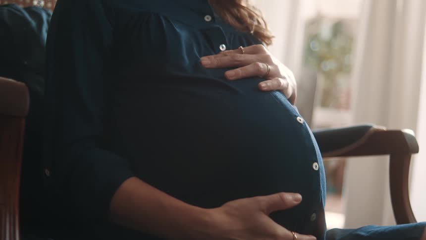 Very pregnant woman sitting on a chair indoors holding her belly