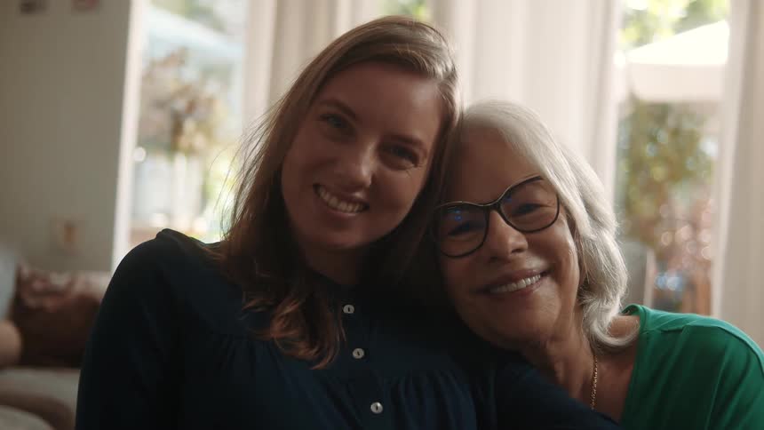 An older mom rests her head on her daughters shoulder as they both smile lovingly for the camera