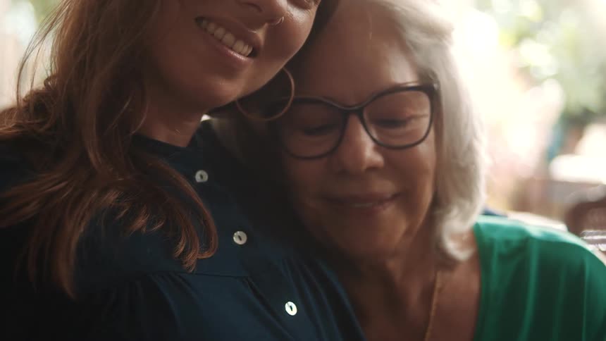 An elderly woman smiles warmly while leaning her head on a younger woman shoulder