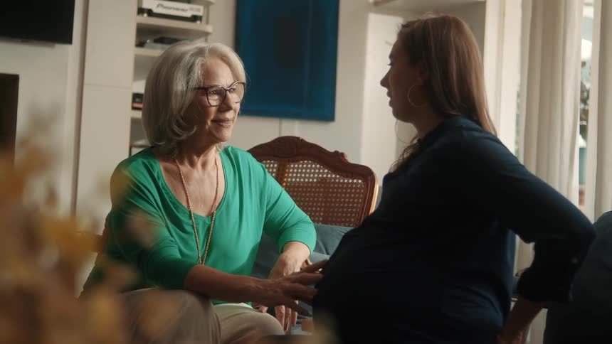 An elderly woman with gray hair and glasses in a green shirt sits facing a pregnant woman with brown hair in a dark blue shirt and the elderly woman touches the pregnant womans stomach