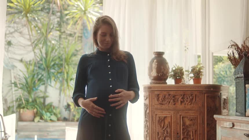 A pregnant woman gently touches her belly while standing in a sunlit cozy indoor space