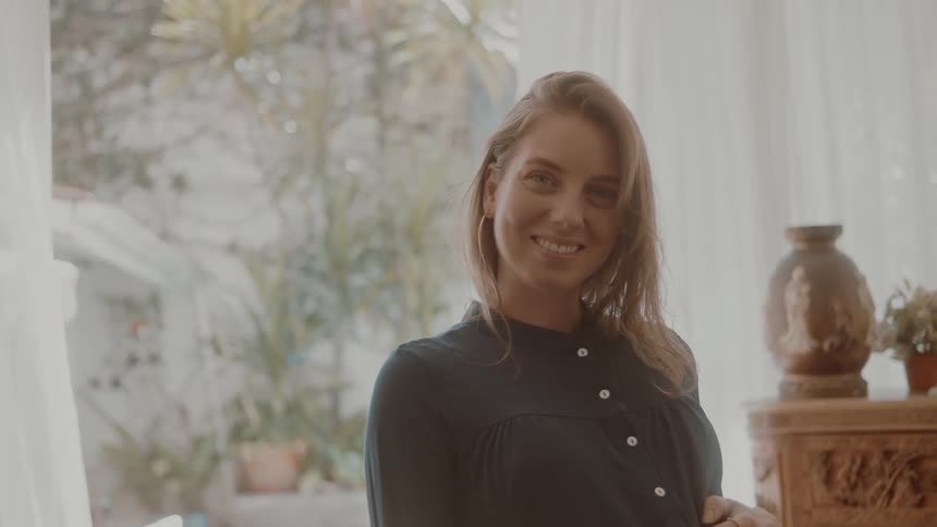 A pregnant woman stands indoors smiling at the camera and gently holding her belly