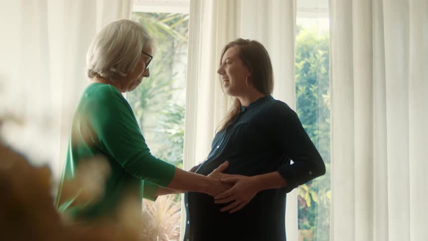 Pregnant woman smiles as older woman touches her belly indoors