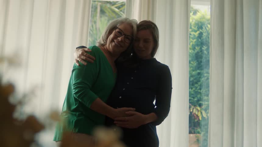 An older woman lovingly embraces a pregnant younger woman near a sunlit window