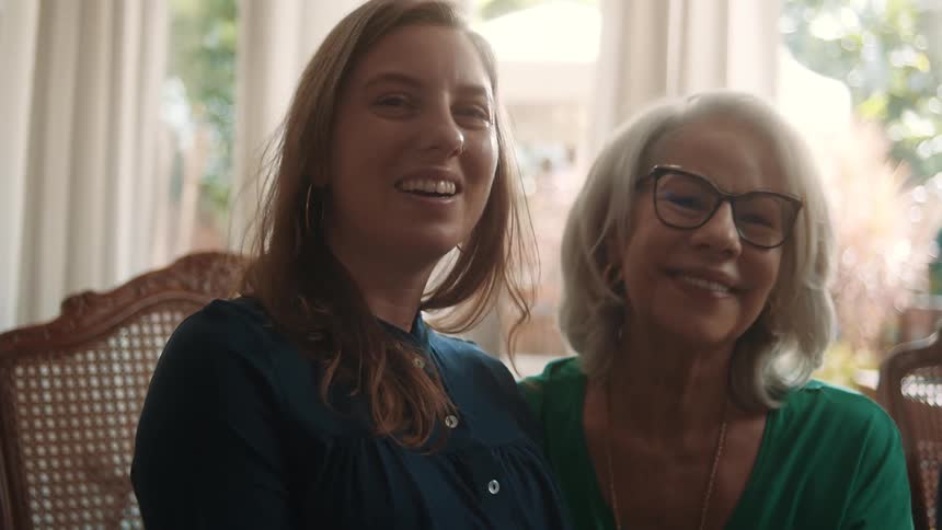 Two women sit closely smiling warmly at each other in a sunlit indoor setting