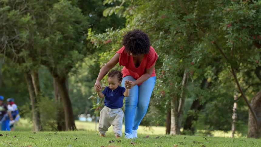 A mother helps her toddler walk through a grassy park