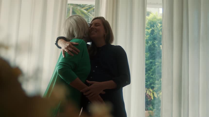 A mother warmly hugs and kisses her pregnant daughter in front of a sunlit window