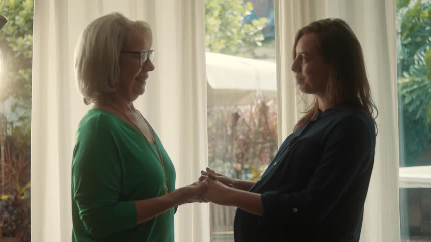 Pregnant woman holds hands with older woman in sunlit home