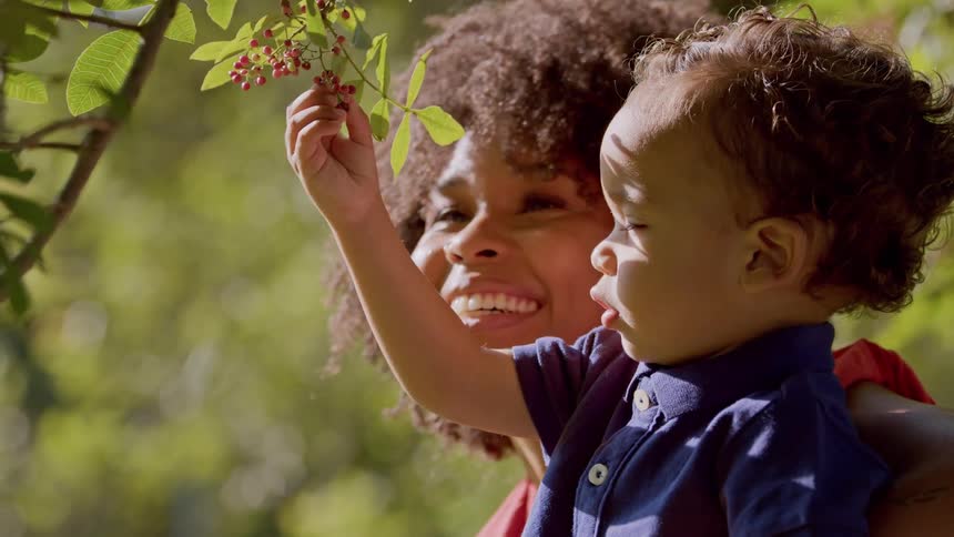 A smiling mom holds her little boy so he can reach out and curiously touch the red berries on a branch