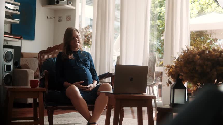 A pregnant woman sits comfortably indoors touching her belly while facing a laptop