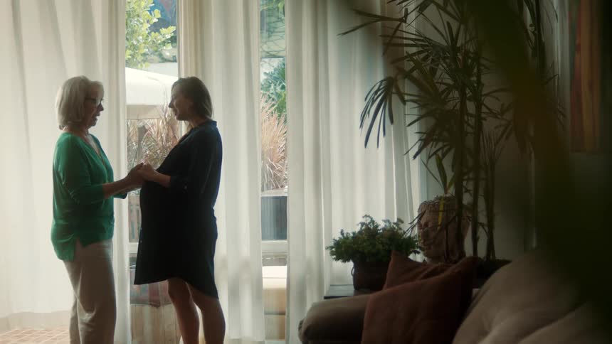 Two women look at each other holding hands in a room with white curtains and plants