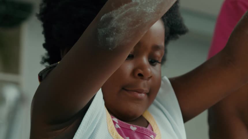 Close up of a childs face as she sprinkles flour onto a surface with flour dusted arms cooking beside an adult in the kitchen