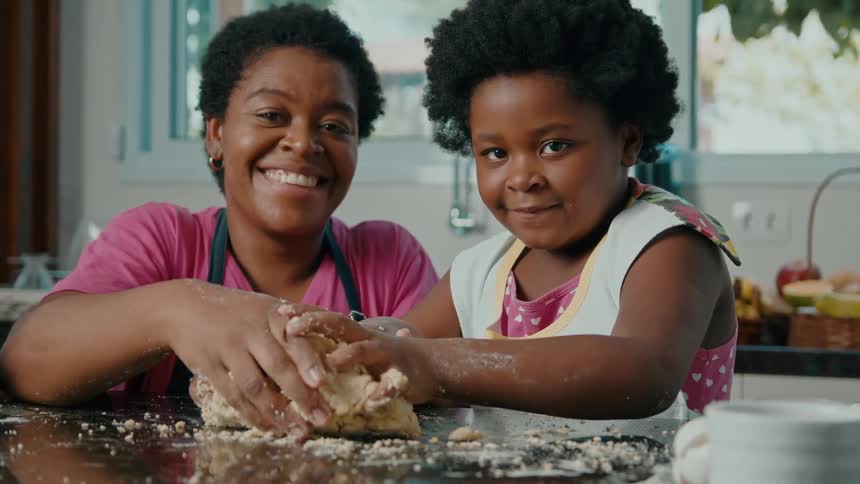 A smiling mother and daughter are making dough together at the kitchen counter
