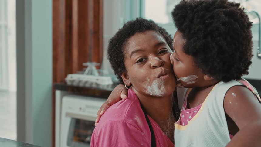 A young child kisses a smiling woman on the cheek in a kitchen both with flour on their faces