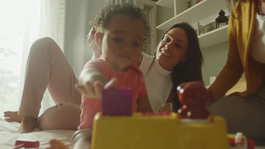 Mother tousles babys hair while baby plays with a toy on the bed